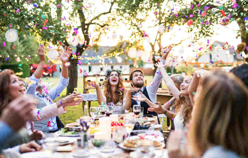 A bride and groom laugh as guests throw colorful confetti at their outdoor wedding venue