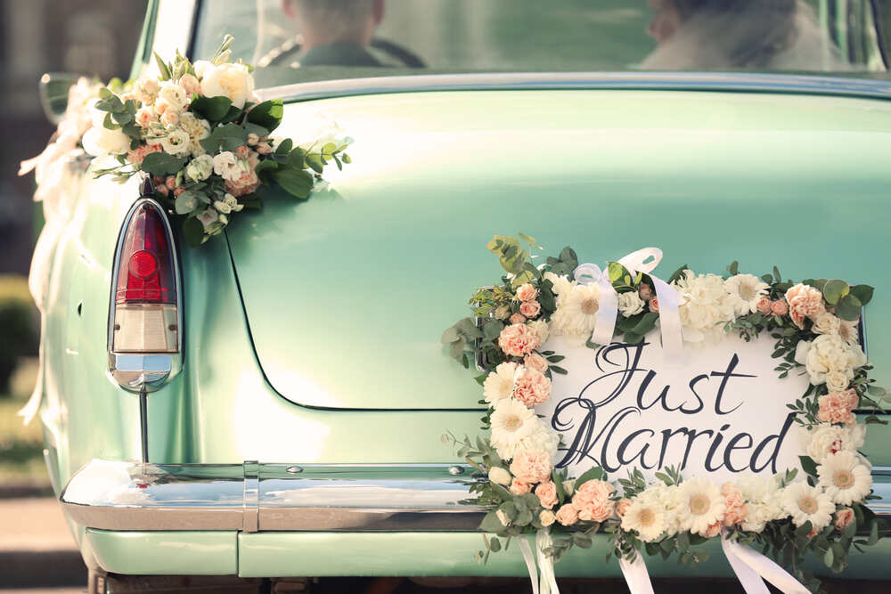 A couple drives away in a classic car with “just married” on the rear bumper 